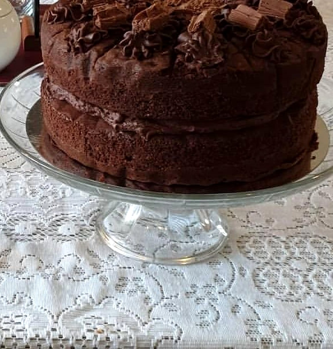 A tall chocolate layer cake topped with chocolate curls on a glass cake stand, over a white lace tablecloth