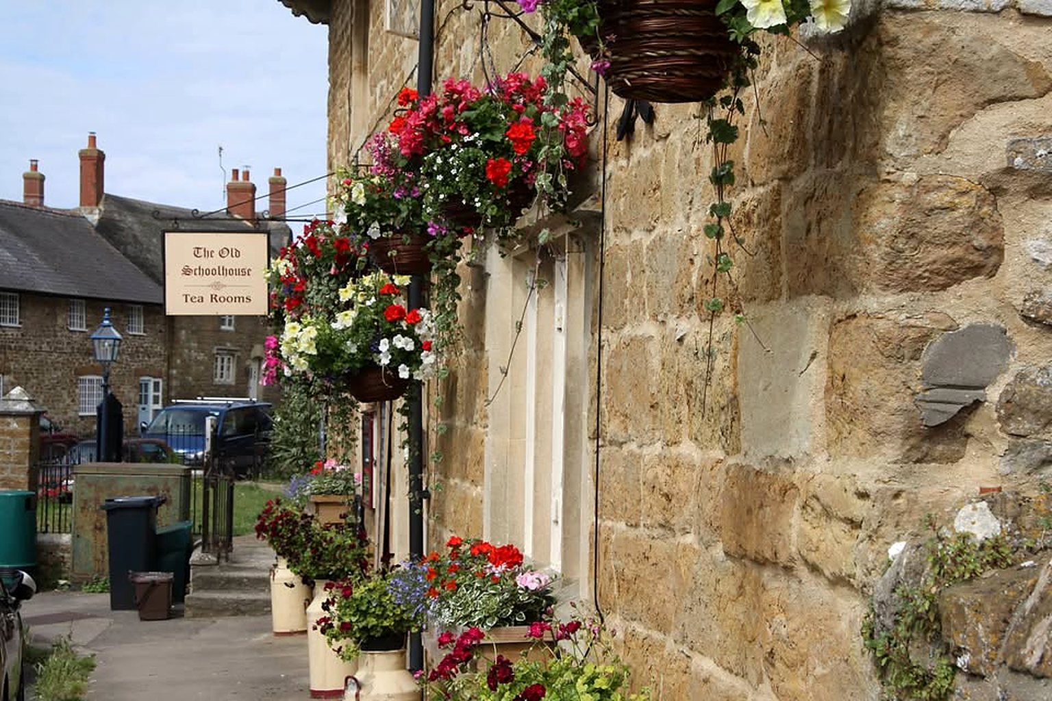 The stone front of the Old Schoolhouse on Back Street, with hanging baskets of red flowers, pots by the door and a small hanging sign reading The Old Schoolhouse Tea Rooms